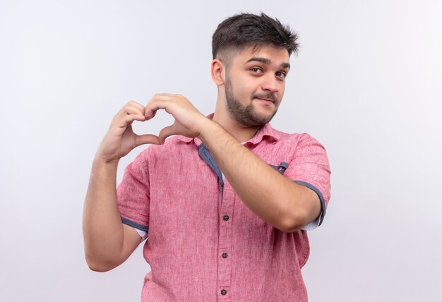 Junger hübscher Kerl, der rosa Poloshirt trägt, das süß schaut, das Liebe singh mit den Händen tut, die über weißer Wand stehen