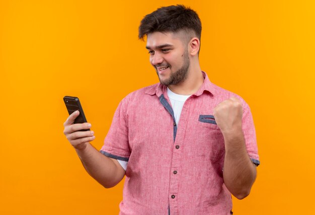 Junger hübscher Kerl, der rosa Poloshirt trägt, das Telefon glücklich tut, tat es mit Faust, die über orange Wand steht