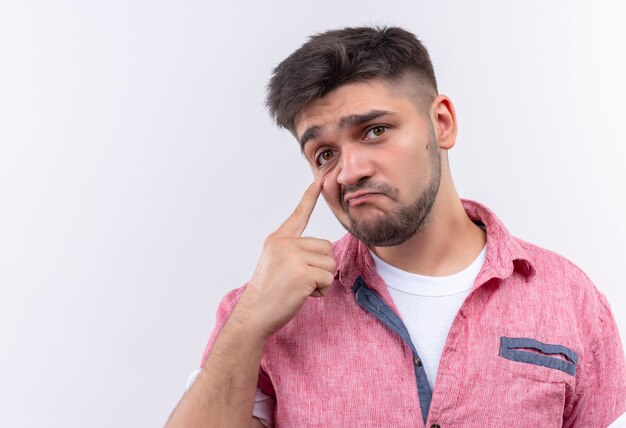 Junger hübscher Kerl, der rosa Poloshirt trägt, das traurig schaut, über weißer Wand stehend zu weinen