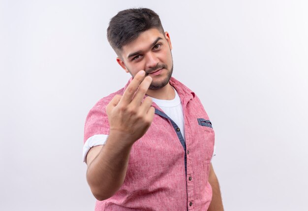 Junger hübscher Kerl, der rosa Poloshirt trägt, das tut, ich beobachte, wie Sie mit den Fingern unterschreiben, die über weißer Wand stehen