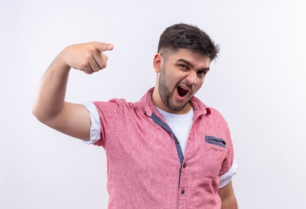 Junger hübscher Kerl, der rosa Poloshirt trägt, das wütend mit Zeigefinger zeigt, der über weißer Wand steht