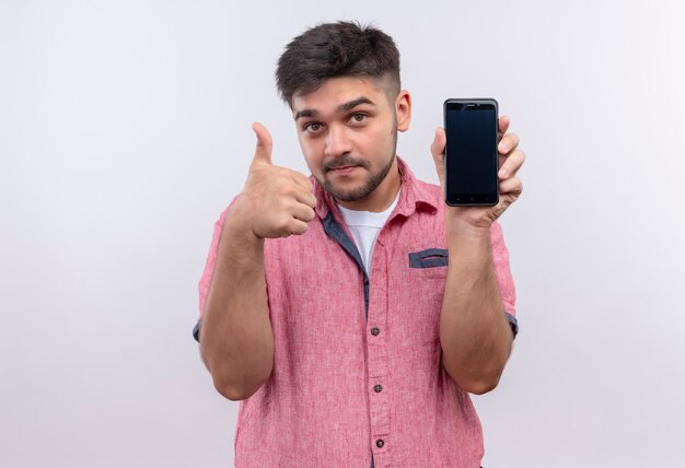 Junger hübscher Kerl, der rosa Poloshirt trägt und das Telefon lobt, das Superzeichen mit Daumen tut, der über weißer Wand steht