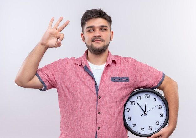 Junger hübscher Kerl, der rosa Poloshirt trägt und Uhr hält, die ok Zeichen mit Finger über weißer Wand steht