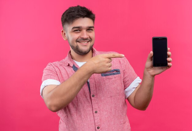 Junger hübscher Kerl, der rosa Poloshirt trägt, zeigt glücklich auf Telefon, das über rosa Wand steht