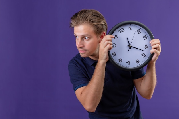 Junger hübscher Kerl, der Uhr hält, die beiseite mit sicherem Ausdruck steht, der über lila Hintergrund steht