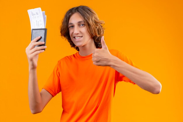 Junger hübscher Kerl im orangefarbenen T-Shirt, das Flugtickets hält, die zuversichtlich zeigen, Daumen hoch lächelnd zu zeigen, die fröhlich über gelbem Hintergrund stehen