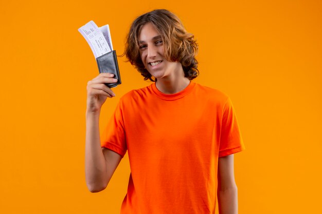 Junger hübscher Kerl im orangefarbenen T-Shirt, das Flugtickets hält Kamera mit dem sicheren Lächeln stehend