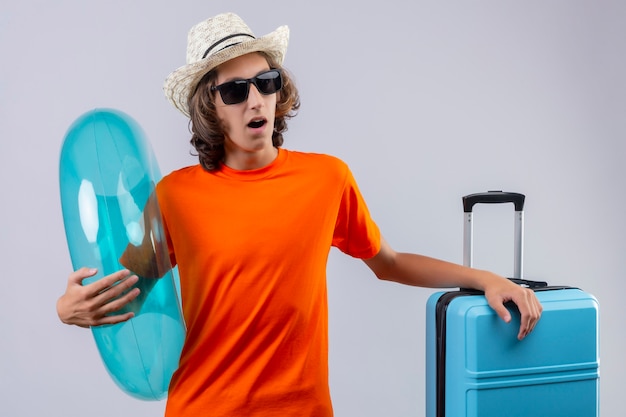 Junger hübscher Kerl im orangefarbenen T-Shirt, das schwarze Sonnenbrille hält, die aufblasbaren Ring hält, der überrascht steht, mit Reisekoffer stehend