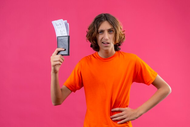Junger hübscher Kerl im orangefarbenen T-Shirt, der Flugtickets hält, die Flugtickets unzufrieden stehen, die über rosa Hintergrund stehen