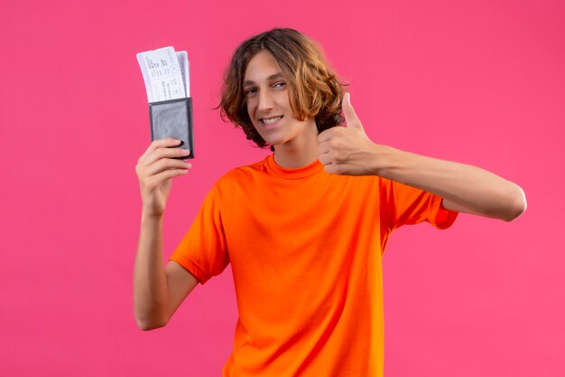 Junger hübscher Kerl im orangefarbenen T-Shirt, der Flugtickets hält, die zuversichtlich zeigen Daumen zeigen, die fröhlich stehend lächeln
