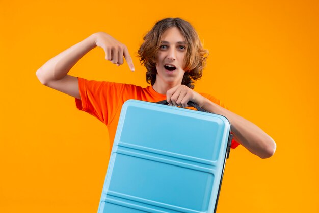 Junger hübscher Kerl im orangefarbenen T-Shirt, der mit dem Reisekoffer steht, der mit dem Finger darauf zeigt, der zuversichtlich und glücklich über gelbem Hintergrund schaut