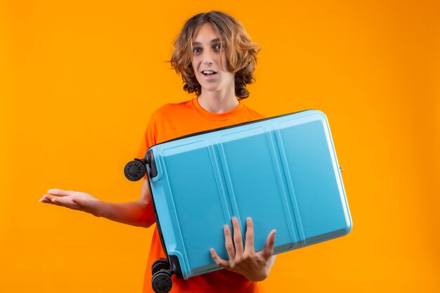 Junger hübscher Kerl im orangefarbenen T-Shirt, der Reisekoffer ahnungslos und verwirrt hält und keine Antwort hat, die Hände verbreitet, die über gelbem Hintergrund stehen