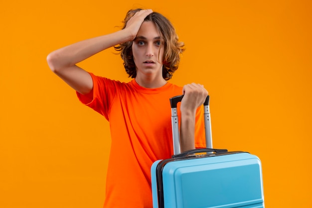 Junger hübscher Kerl im orangefarbenen T-Shirt, der Reisekoffer hält, der mit Hand auf Kopf für Fehler steht, der verwirrt schaut, erinnert sich Fehler über gelben Hintergrund