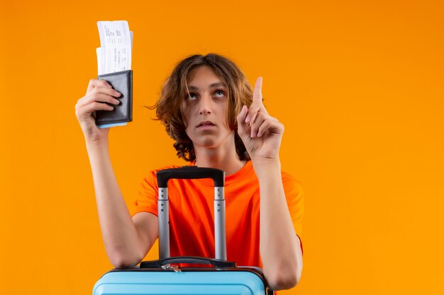 Junger hübscher Kerl im orangefarbenen T-Shirt, der Reisekoffer und Flugtickets hält, die oben schauen und auf etwas stehendes zeigen