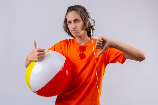 Junger hübscher Kerl im orangefarbenen T-Shirt mit Kopfhörern, die aufblasbaren Ball halten, missfiel, Daumen hoch und unten stehend über weißem Hintergrund zeigend