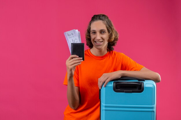 Junger hübscher Kerl im orangefarbenen T-Shirt stehend mit Reisekoffer, der Flugtickets betrachtet, die Kamera glücklich und positiv lächelnd betrachten