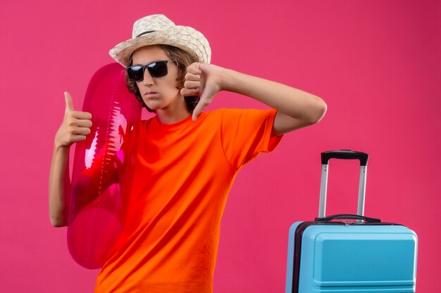 Junger hübscher Kerl im orangefarbenen T-Shirt und im Sommerhut, der schwarze Sonnenbrille hält, die aufblasbaren Ring hält, missfiel zeigt Daumen hoch und runter mit negativem Ausdruck auf Gesicht, das mit Reisen steht