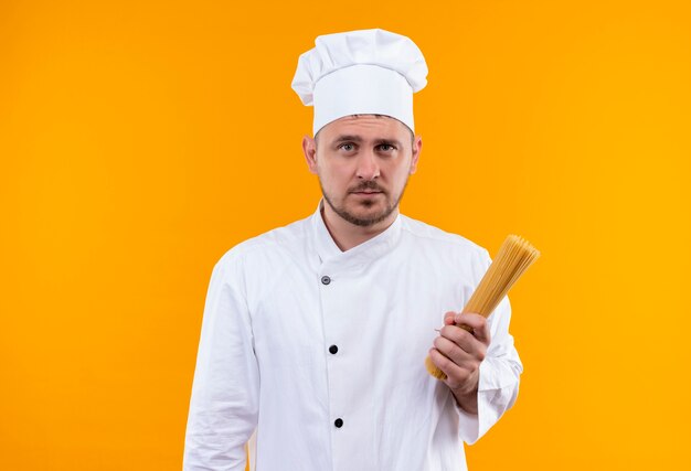 Junger hübscher Koch in der Kochuniform, die Spaghetti-Nudeln hält, die auf orange Raum lokalisiert suchen