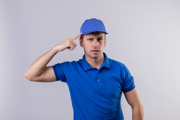 Junger hübscher Lieferbote in blauer Uniform und Mütze, die seine Schläfe mit dem Finger zeigen