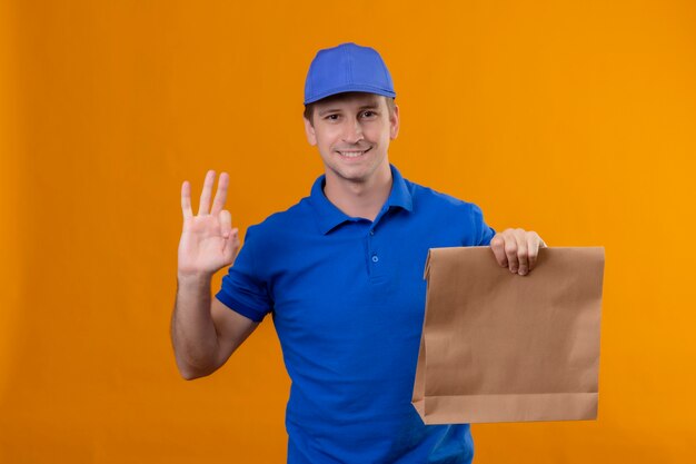 Junger hübscher Lieferbote in der blauen Uniform und in der Kappe, die Papierpaket positiv und glücklich hält, ok Zeichen, das über orange Wand steht
