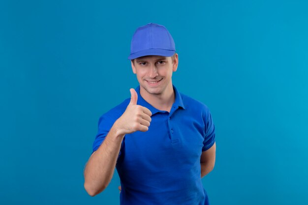 Junger hübscher Lieferbote in der blauen Uniform und in der Kappe glücklich und positiv lächelnd freundlich zeigt Daumen hoch stehend über blauer Wand