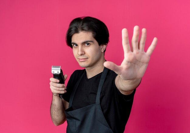 junger hübscher männlicher Friseur in der Uniform, die Haarschneidemaschinen hält und Hand lokalisiert auf rosa Wand hält