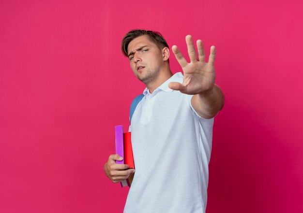 junger hübscher männlicher Student, der Rückentasche hält, die Hand lokalisiert auf rosa Wand hält