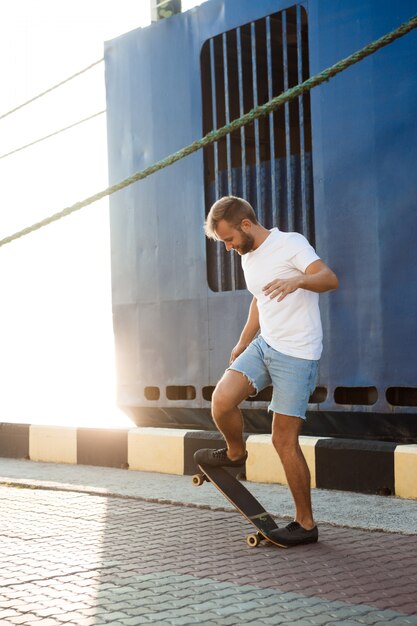 Junger hübscher Mann, der am Meer geht, lächelnd, Skateboarding.