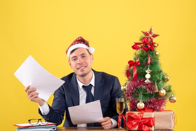 junger hübscher Mann, der am Tisch nahe Weihnachtsbaum sitzt und auf Gelb präsentiert