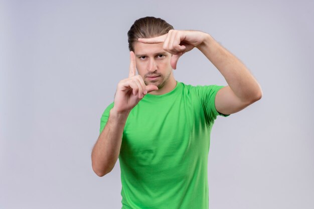 Junger hübscher Mann, der grünes T-Shirt trägt, das durch einen Rahmen schaut, der durch seine Hände gebildet wird