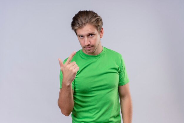 Junger hübscher Mann, der grünes T-Shirt trägt, das mit dem Finger auf sich selbst mit dem selbstbewussten Ausdruck zeigt, der über weißer Wand steht