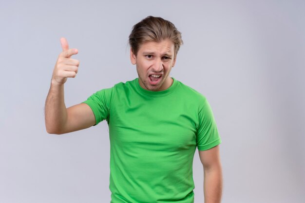 Junger hübscher Mann, der grünes T-Shirt trägt, das mit dem Finger zur Kamera zeigt, missfiel das Streiten, das über weißer Wand steht