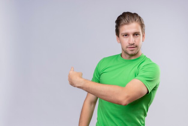 Junger hübscher Mann, der grünes T-Shirt trägt, das schaut, lächelt und auf etwas hinter zeigt