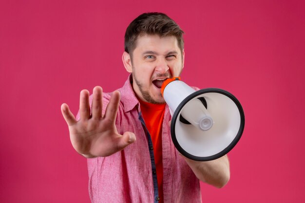 Junger hübscher Mann, der Megaphon hält, das zu ihm schreit und Stoppschild mit Hand macht