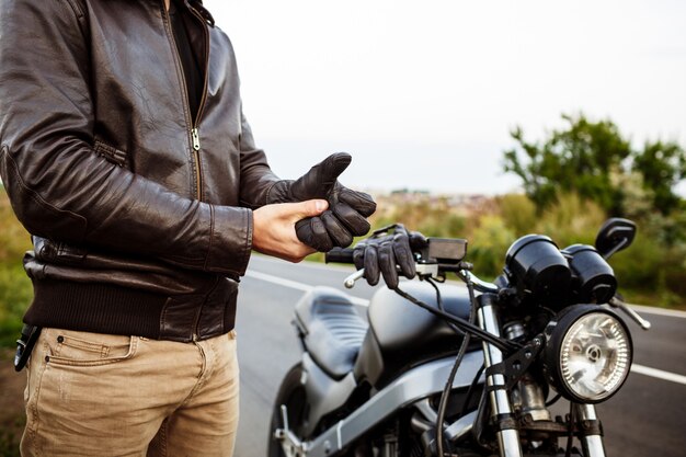 Junger hübscher Mann, der nahe seinem Motorrad aufwirft, Handschuhe tragend.