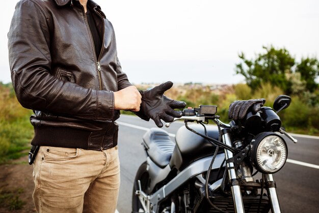 Junger hübscher Mann, der nahe seinem Motorrad aufwirft, Handschuhe tragend.