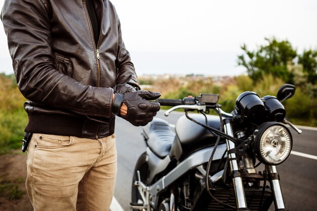 Junger hübscher Mann, der nahe seinem Motorrad aufwirft, Handschuhe tragend.