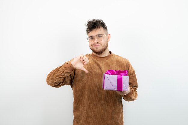 Junger hübscher Mann, der sein Feiertagsgeschenk hält und Daumen unten zeigt.