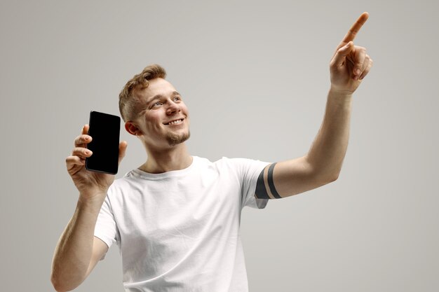 Junger hübscher Mann, der Smartphonebildschirm über grauem Hintergrund mit einem Überraschungsgesicht zeigt. Menschliche Emotionen, Gesichtsausdruckkonzept