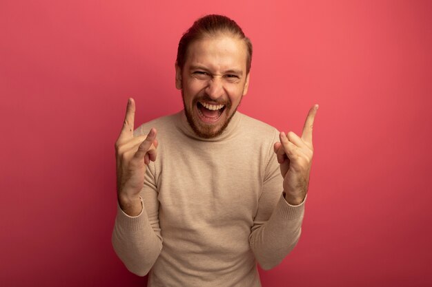 Junger hübscher Mann im beigen Rollkragenpullover verrückt glücklich, das Steinsymbol macht, das über rosa Wand steht