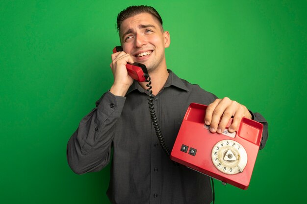 Junger hübscher Mann im grauen Hemd, das auf Weinlese-Telefon spricht, das fröhlich über grüner Wand steht