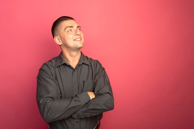 Junger hübscher Mann im grauen Hemd, das beiseite mit Lächeln auf Gesicht mit verschränkten Armen auf seiner Brust schaut, die über rosa Wand stehen