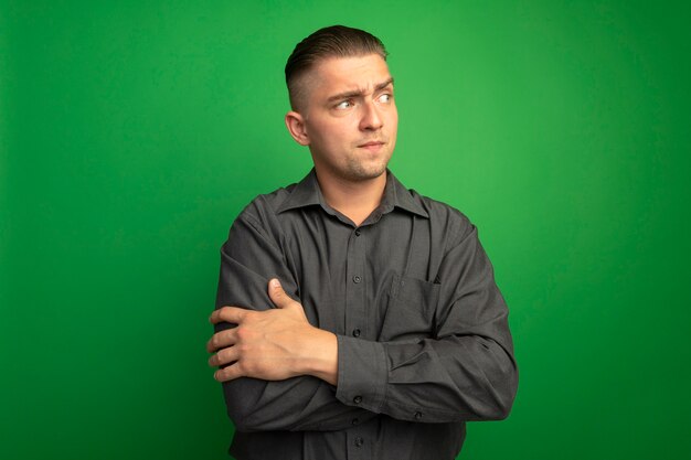 Junger hübscher Mann im grauen Hemd, das beiseite mit skeptischem Ausdruck steht, der über grüner Wand steht