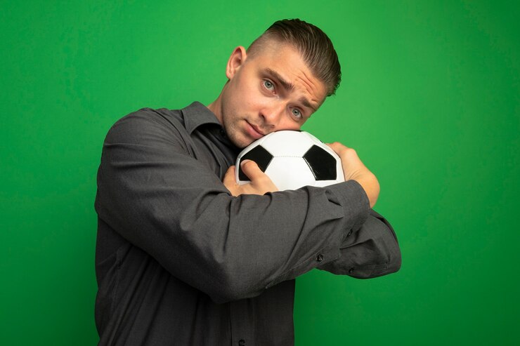 Kostenlose Foto junger hübscher mann im grauen hemd, das fußball umarmt, der seinen kopf auf ball lehnt, der vorne mit traurigem ausdruck steht, der über grüner wand steht
