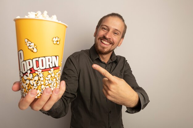 Junger hübscher Mann im grauen Hemd, der Eimer mit Popcorn zeigt, der mit Indexfigner darauf zeigt, der fröhlich lächelt