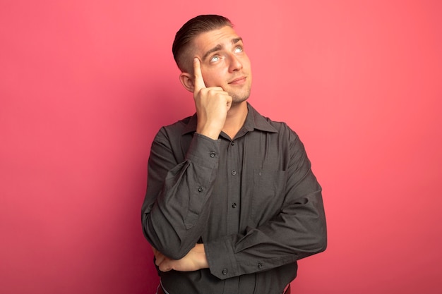 Junger hübscher Mann im grauen Hemd, der oben mit dem Finger auf seinem Gesicht denkt und positiv steht über rosa Wand