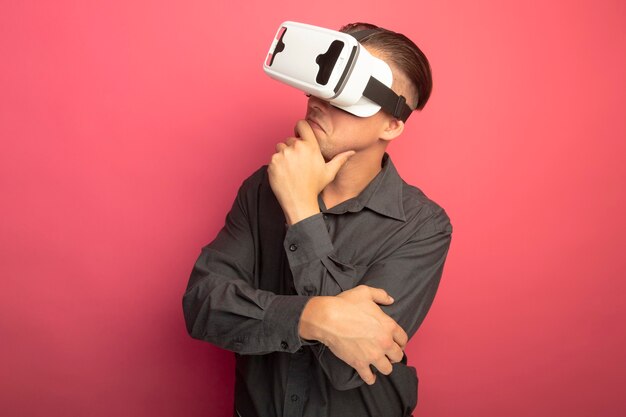 Junger hübscher Mann im grauen Hemd mit der Brille der virtuellen Realität, die mit Händen gestikuliert, die mit Hand auf Kinn beiseite schauen