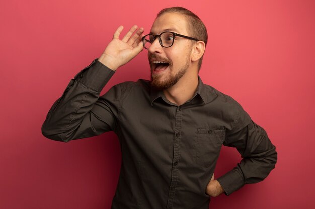 Junger hübscher Mann im grauen Hemd und in den Gläsern, die glücklich und positiv beiseite schauen und seine Brille berühren