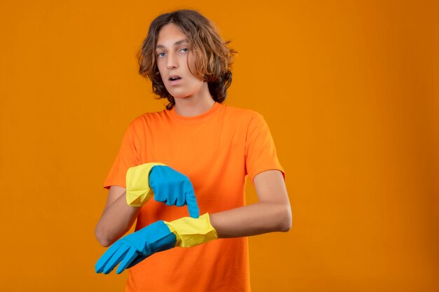 Junger hübscher Mann im orangefarbenen T-Shirt mit Gummihandschuhen, die auf seine Hand zeigen, die an die Zeit erinnert, die Kamera überrascht und ängstlich betrachtet, die über gelbem Hintergrund stehen
