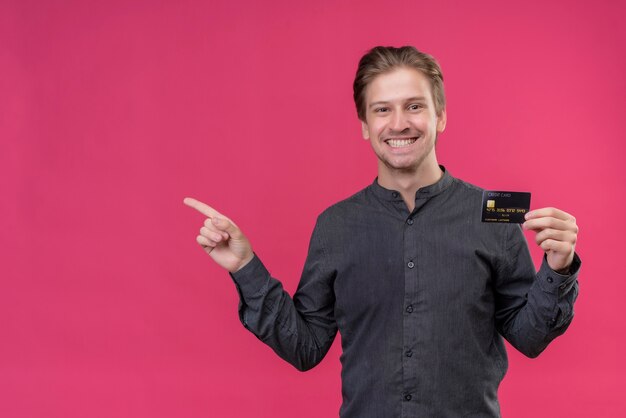 Junger hübscher Mann im schwarzen Hemd, das Kreditkarte hält und mit dem Finger zur Seite zeigt